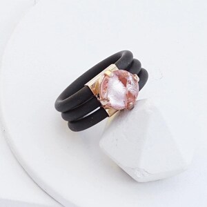 May include: A black silicone ring with three bands and a rose gold setting with a pink cracked glass stone.