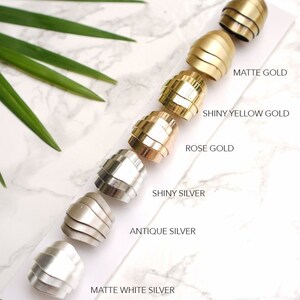 May include: A close-up of seven metal samples in different finishes, including matte gold, shiny yellow gold, rose gold, shiny silver, antique silver, and matte white silver. The samples are arranged in a row from left to right, with the matte gold sample at the top and the matte white silver sample at the bottom. The samples are all the same shape and size, and they are all made of a smooth, shiny metal.