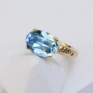 May include: A ring featuring a large, oval-cut, light blue aquamarine gemstone set in a textured, shiny yellow gold band. The text AQUAMARINE | SHINY YELLOW GOLD is printed at the bottom of the image, describing the stone and metal finish.