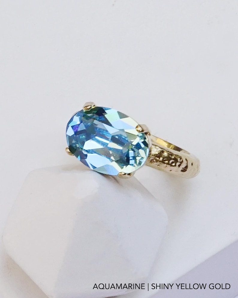 May include: A ring featuring a large, oval-cut, light blue aquamarine gemstone set in a textured, shiny yellow gold band. The text AQUAMARINE | SHINY YELLOW GOLD is printed at the bottom of the image, describing the stone and metal finish.