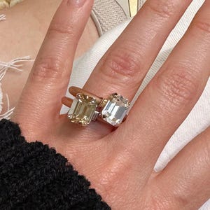 Op de afbeelding: Een dubbele ring met twee rechthoekige stenen in smaragdgeslepen vorm. De ene steen heeft een lichtgele tint, terwijl de andere een heldere, kleurloze steen is. De ringbanden zijn van metaal, waarbij de ene roségoud en de andere zilverkleurig is.