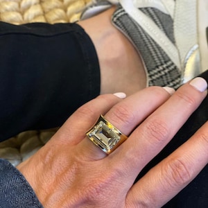 May include: A wide gold band ring featuring a large, rectangular, emerald-cut clear gemstone set in the center. The metal band has a polished, reflective finish and a slightly concave shape that tapers toward the sides of the finger.