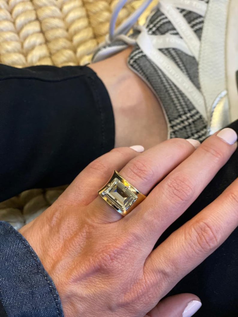 May include: A wide gold band ring featuring a large, rectangular, emerald-cut clear gemstone set in the center. The metal band has a polished, reflective finish and a slightly concave shape that tapers toward the sides of the finger.