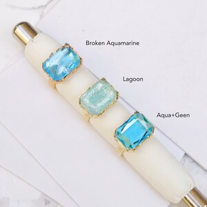 May include: Three gold rings with different colored glass stones. The stones are labeled "Broken Aquamarine", "Lagoon", and "Aqua+Green".