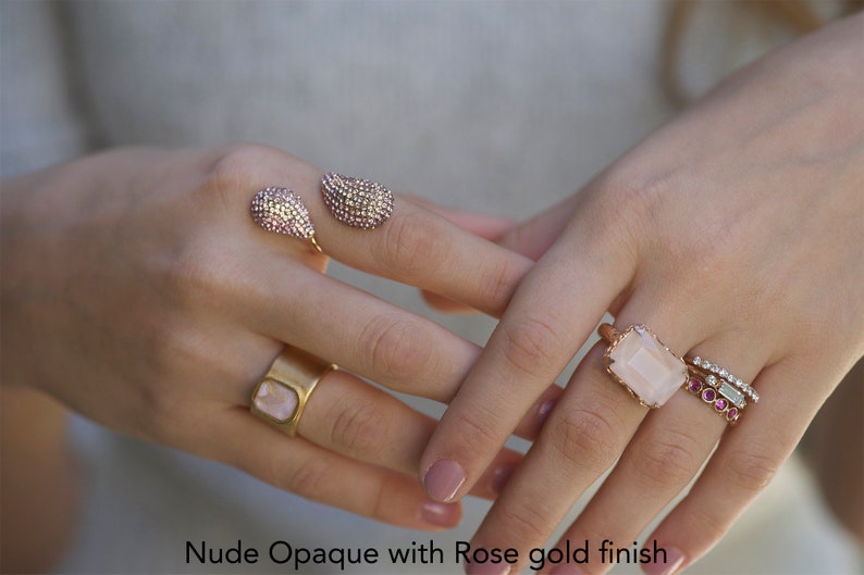 May include: A set of four rings with rose gold finish. The rings feature a variety of stones, including a large rectangular pink stone, smaller pink stones, and clear stones. The text "Nude Opaque with Rose gold finish" is visible at the bottom of the image.