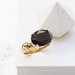 May include: A gold ring featuring a large, oval-cut black gemstone set in a four-prong mounting. The band of the ring has a textured, hammered finish that reflects light across its surface. The word Black is printed in the lower left corner.