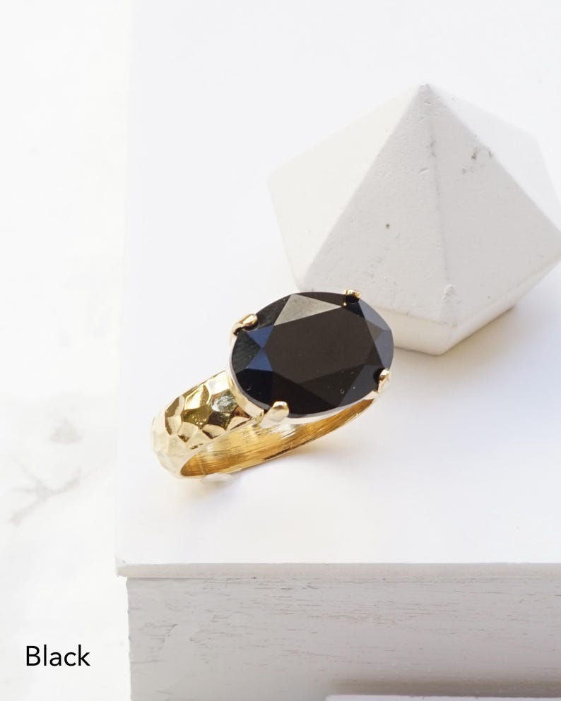 May include: A gold ring featuring a large, oval-cut black gemstone set in a four-prong mounting. The band of the ring has a textured, hammered finish that reflects light across its surface. The word Black is printed in the lower left corner.
