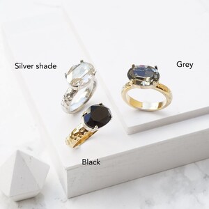 May include: Three rings with oval-cut stones in silver shade, grey, and black. The silver shade ring has a textured silver band, the grey ring has a gold band, and the black ring has a textured gold band. Each ring is labeled with its respective color.