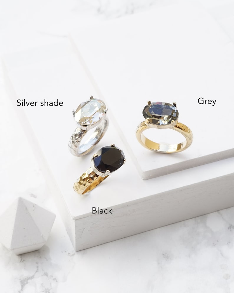 May include: Three rings with oval-cut stones in silver shade, grey, and black. The silver shade ring has a textured silver band, the grey ring has a gold band, and the black ring has a textured gold band. Each ring is labeled with its respective color.
