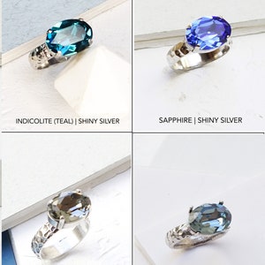 May include: Four rings with oval-cut faceted stones in teal, blue, grey, and light blue. Each ring features a textured, hammered-style metal band. The text labels indicate the colors as Indicolite, Sapphire, Grey, and Indian Sapphire with silver finishes.