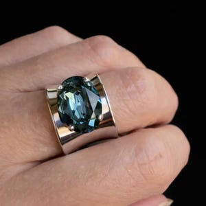 May include: A wide silver-toned band ring featuring a large, oval-cut, dark teal-coloured gemstone set in the centre. The gemstone has multiple facets that reflect light, and the metal band has a smooth, polished finish with a curved design.