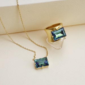 May include: A gold chain necklace and ring with a blue-green rectangular gemstone. The gemstone is labeled "Erinite (Swarovski)".
