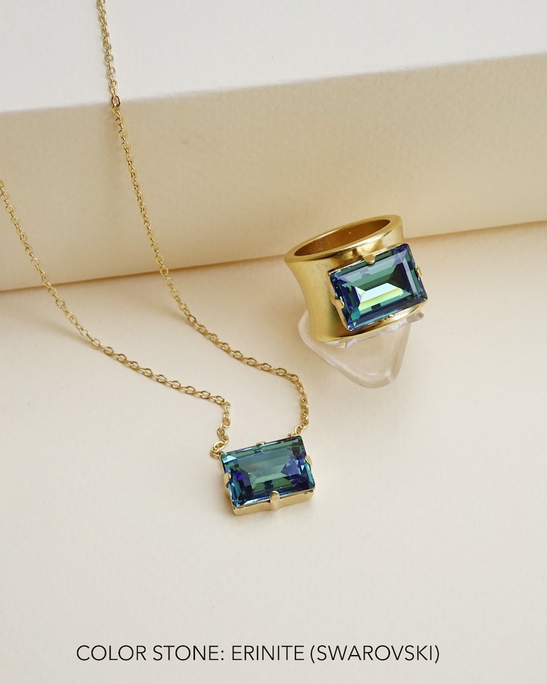 May include: A gold chain necklace and ring with a blue-green rectangular gemstone. The gemstone is labeled "Erinite (Swarovski)".