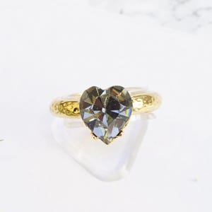 May include: A ring featuring a faceted gray heart-shaped stone set on a band with a shiny yellow gold finish. The band has a textured, hammered surface. The text "Gray with shiny yellow gold finish" is printed at the bottom of the image.