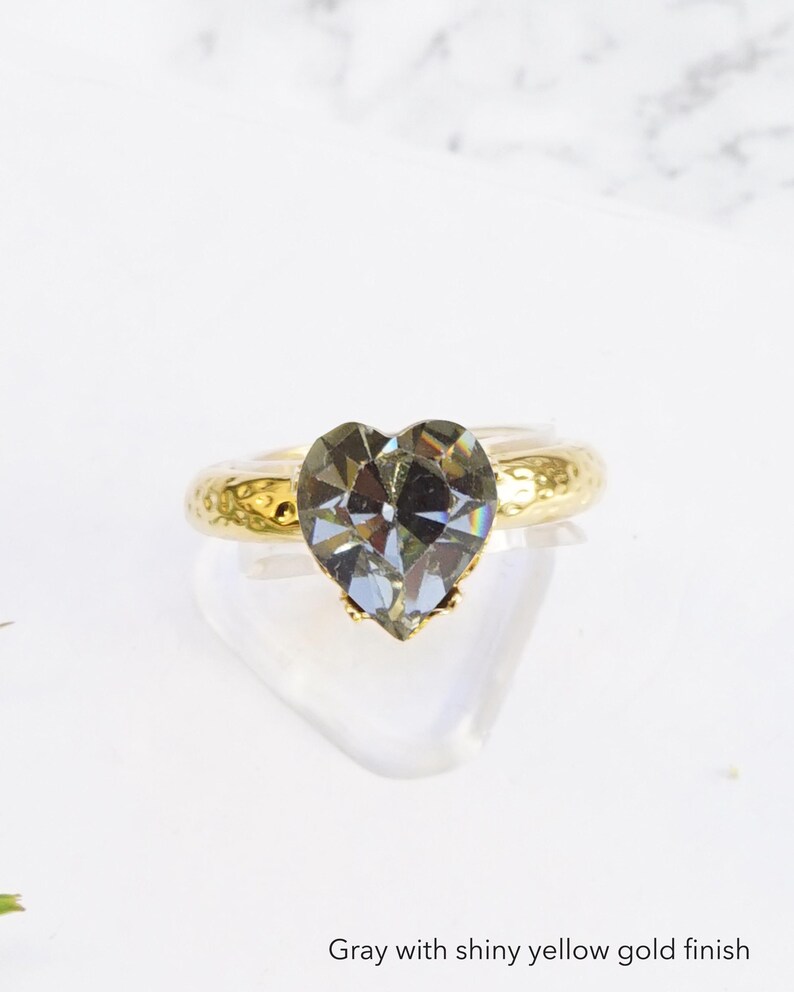May include: A ring featuring a faceted gray heart-shaped stone set on a band with a shiny yellow gold finish. The band has a textured, hammered surface. The text "Gray with shiny yellow gold finish" is printed at the bottom of the image.