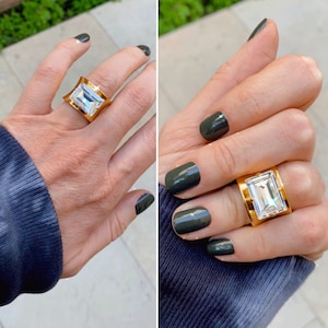May include: A wide gold band ring featuring a large, rectangular, emerald-cut clear stone set in the center. The metal band has a polished finish and a slightly concave shape that tapers toward the finger, showcasing a bright, reflective surface.