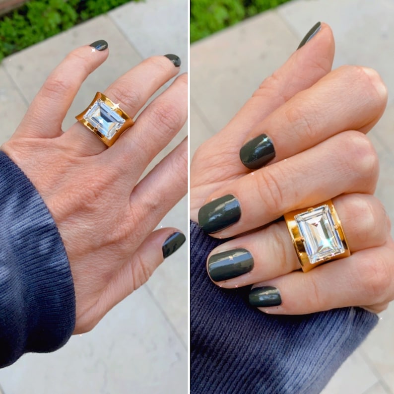 May include: A wide gold band ring featuring a large, rectangular, emerald-cut clear stone set in the center. The metal band has a polished finish and a slightly concave shape that tapers toward the finger, showcasing a bright, reflective surface.