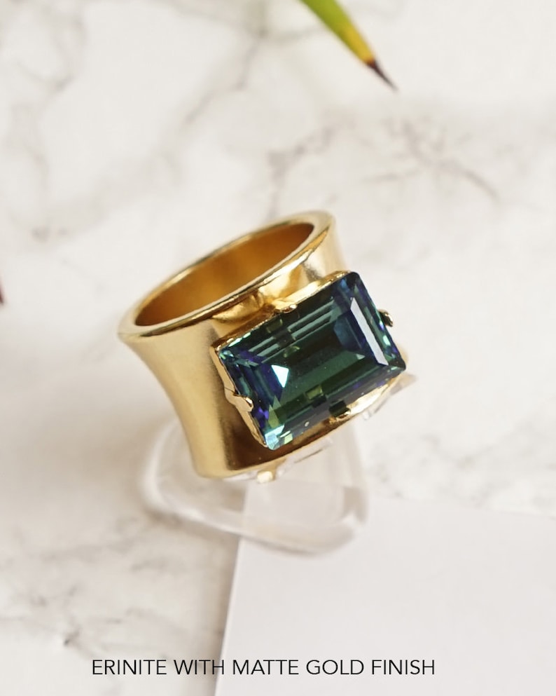 May include: A wide band ring featuring a matte gold finish and a large, rectangular, emerald-cut erinite gemstone. The ring is displayed with the text ERINITE WITH MATTE GOLD FINISH printed at the bottom of the frame.