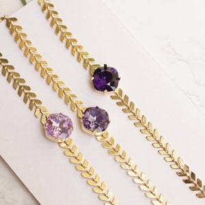 Puede incluir: Tres pulseras doradas con una cadena de patr&oacute;n en espiga y un cristal cuadrado &uacute;nico. Los cristales son de color violeta, tanzanita y purple velvet de izquierda a derecha, seg&uacute;n lo indicado por el texto en la imagen.