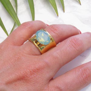 May include: A wide gold-toned ring featuring a central oval-shaped light blue cabochon stone. The band has a smooth, polished finish and a curved design that tapers slightly towards the centre where the stone is securely mounted in a prong setting.