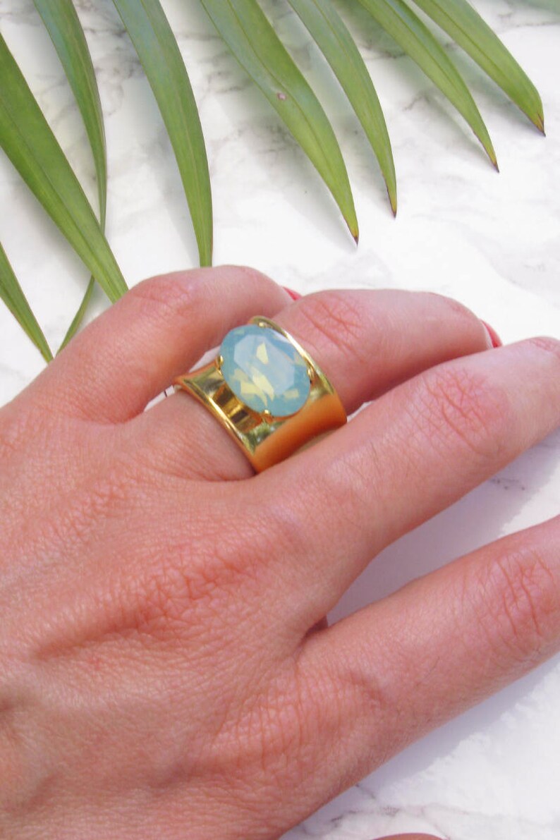 May include: A wide gold-toned ring featuring a central oval-shaped light blue cabochon stone. The band has a smooth, polished finish and a curved design that tapers slightly towards the centre where the stone is securely mounted in a prong setting.