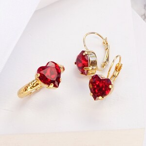 May include: A gold ring and a pair of matching leverback earrings, each featuring a faceted heart-shaped red stone. The metal has a shiny yellow gold finish and a textured band on the ring. The text at the bottom reads SIAM | SHINY YELLOW GOLD.