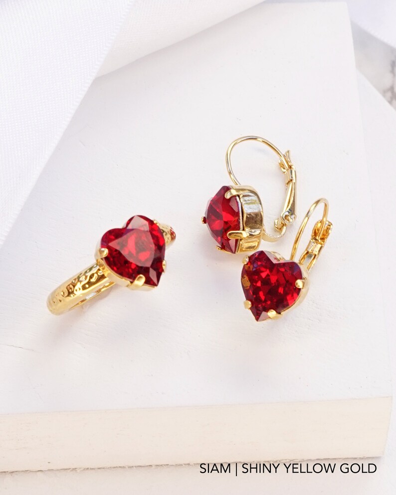 May include: A gold ring and a pair of matching leverback earrings, each featuring a faceted heart-shaped red stone. The metal has a shiny yellow gold finish and a textured band on the ring. The text at the bottom reads SIAM | SHINY YELLOW GOLD.
