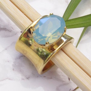 May include: A wide gold-toned metal ring featuring a large, oval-shaped, faceted light blue stone set in a four-prong setting. The ring band has a smooth, polished finish and a curved, open design that tapers slightly towards the centre.