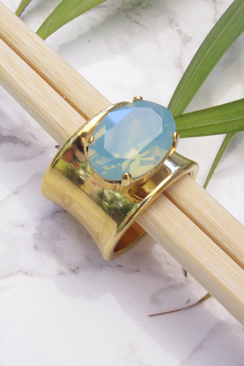 May include: A wide gold-toned metal ring featuring a large, oval-shaped, faceted light blue stone set in a four-prong setting. The ring band has a smooth, polished finish and a curved, open design that tapers slightly towards the centre.