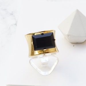 May include: A wide gold-tone ring featuring a large, rectangular black stone set in the center with four small prongs. The band has a smooth, matte gold finish. The text at the bottom of the image reads: BLACK WITH MATTE GOLD FINISH.