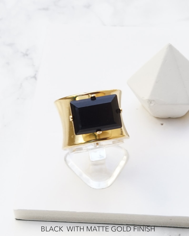 May include: A wide gold-tone ring featuring a large, rectangular black stone set in the center with four small prongs. The band has a smooth, matte gold finish. The text at the bottom of the image reads: BLACK WITH MATTE GOLD FINISH.