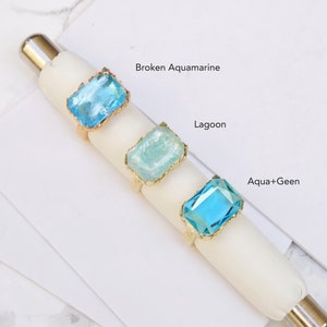 May include: Three gold rings with different colored glass stones. The stones are labeled "Broken Aquamarine", "Lagoon", and "Aqua+Green".