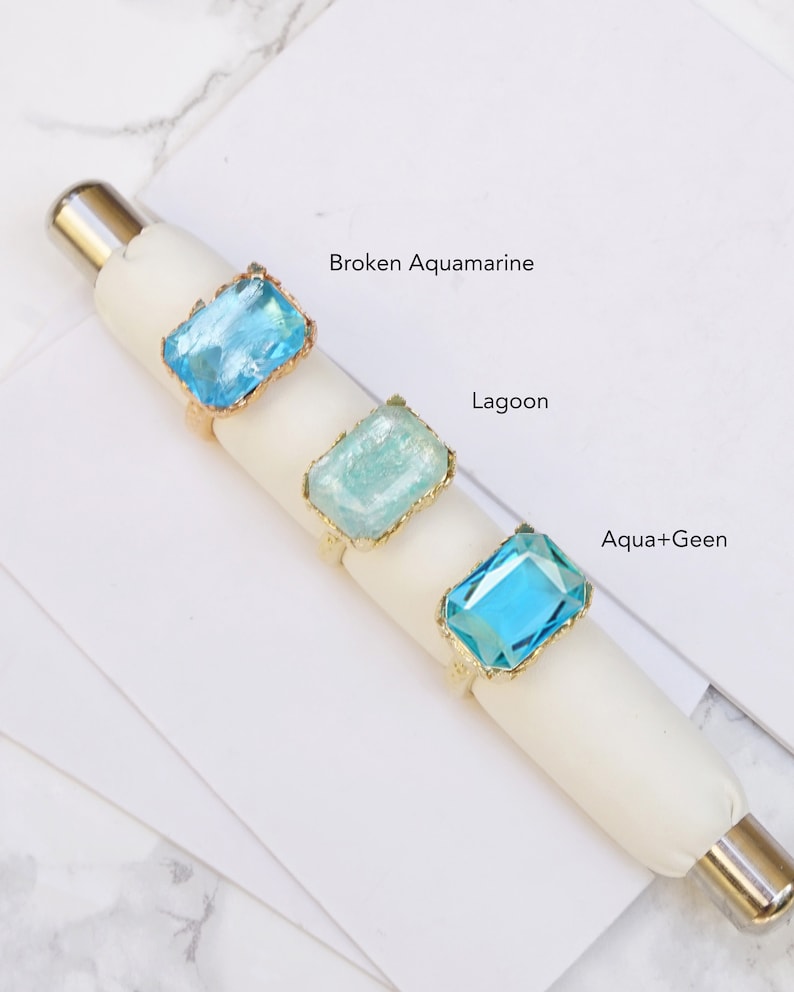 May include: Three gold rings with different colored glass stones. The stones are labeled "Broken Aquamarine", "Lagoon", and "Aqua+Green".