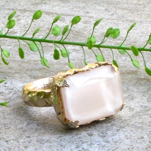 May include: A ring with a large, rectangular, opaque pink stone set in a gold setting with leaf-like details. The ring has a rose gold finish.