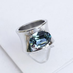 May include: A wide band ring featuring a polished shiny silver finish and a large, oval-shaped Indian sapphire crystal set in the centre. The faceted blue-grey stone is secured by four small prongs, creating a bold and reflective metallic appearance.
