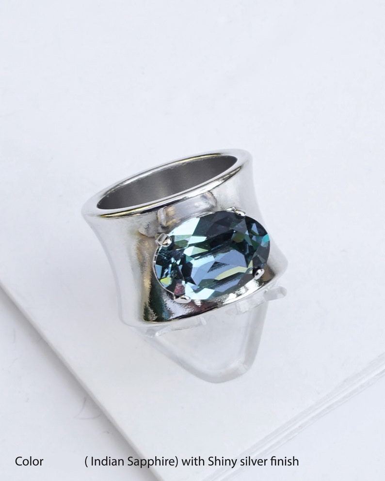 May include: A wide band ring featuring a polished shiny silver finish and a large, oval-shaped Indian sapphire crystal set in the centre. The faceted blue-grey stone is secured by four small prongs, creating a bold and reflective metallic appearance.