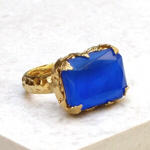 K&ouml;nnte beinhalten: Ein goldener Ring mit einem blauen, undurchsichtigen Edelstein, der in einer blattf&ouml;rmigen Fassung sitzt. Die Ringschiene ist strukturiert.