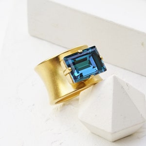May include: A gold ring with a teal blue square gemstone. The ring is matte gold and the gemstone is set in a bezel setting. The ring is on a white surface.