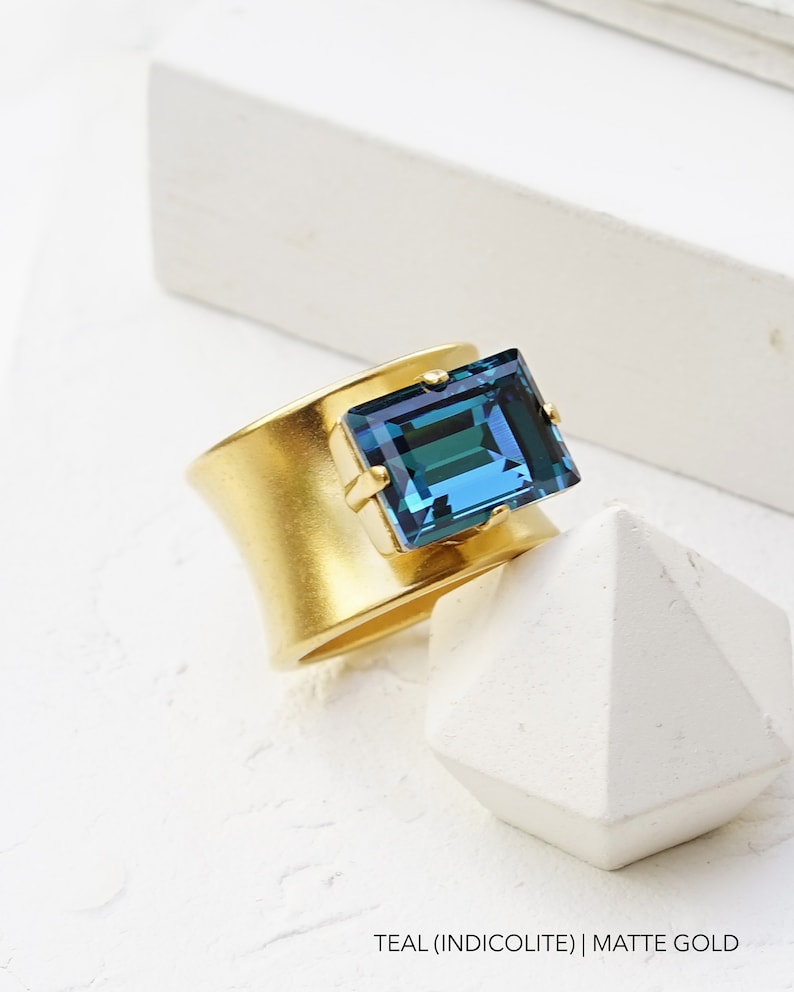 May include: A gold ring with a teal blue square gemstone. The ring is matte gold and the gemstone is set in a bezel setting. The ring is on a white surface.