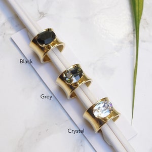 May include: Three gold-toned rings with wide bands, each featuring a large oval-shaped stone. The rings are labelled Black, Grey, and Crystal, corresponding to the black, dark grey, and clear faceted stones set in the centre of each metal band.
