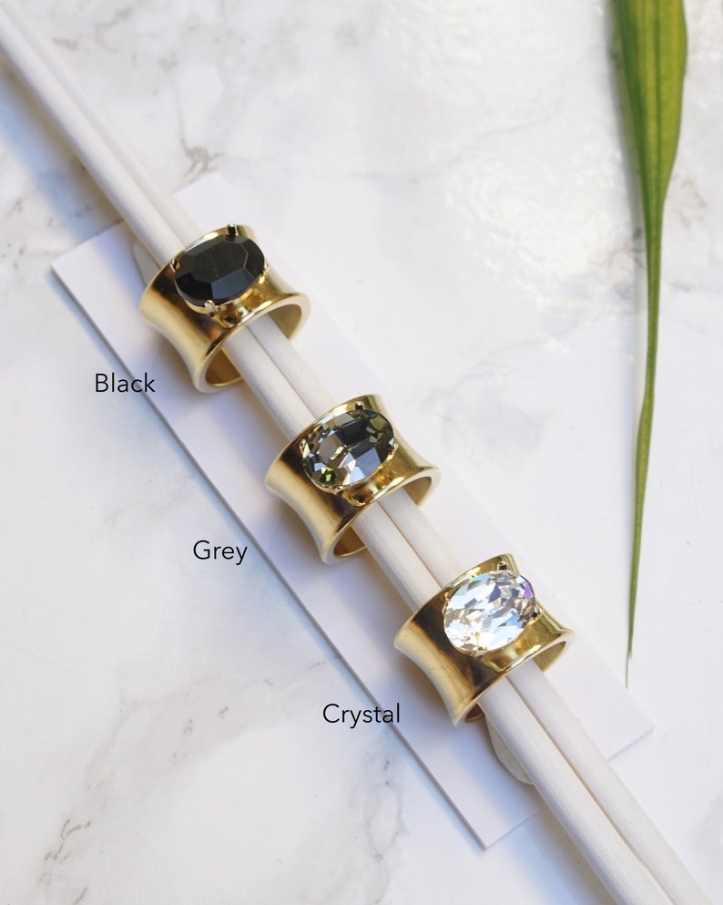 May include: Three gold-toned rings with wide bands, each featuring a large oval-shaped stone. The rings are labelled Black, Grey, and Crystal, corresponding to the black, dark grey, and clear faceted stones set in the centre of each metal band.