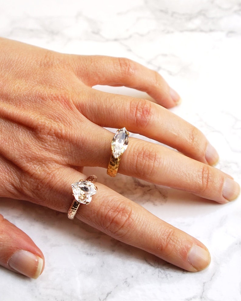 May include: Two rings with textured bands and clear faceted stones. One ring features a heart-shaped stone on a rose-toned band, while the other displays a marquise-cut stone on a yellow-toned band. Both rings have a hammered metal finish.