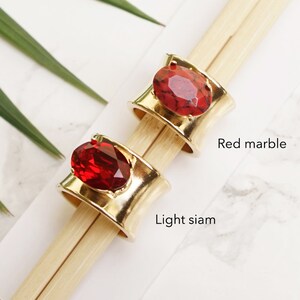 May include: Two wide gold-tone metal rings featuring large oval-shaped stones. One ring is set with a red marble stone labelled Red marble, and the other ring is set with a faceted deep red stone labelled Light siam. The rings are displayed on wooden sticks.