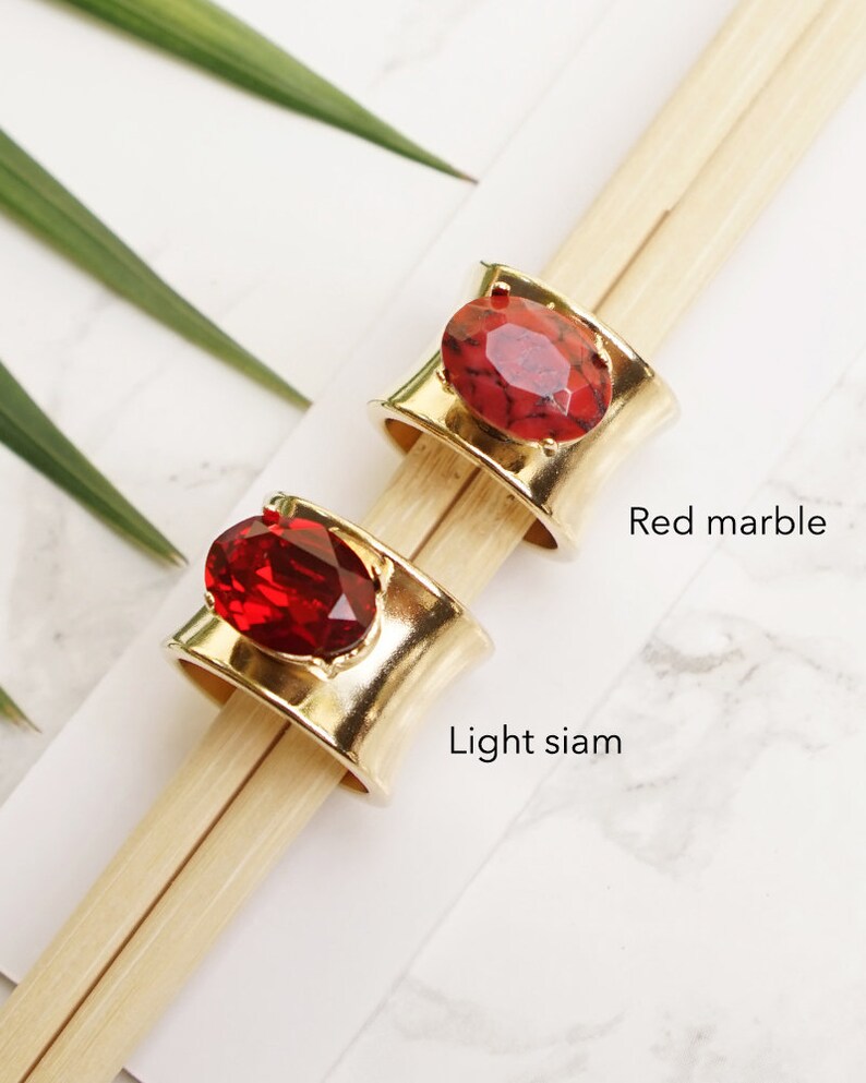 May include: Two wide gold-tone metal rings featuring large oval-shaped stones. One ring is set with a red marble stone labelled Red marble, and the other ring is set with a faceted deep red stone labelled Light siam. The rings are displayed on wooden sticks.