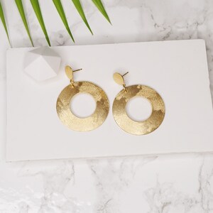 May include: A pair of gold-toned earrings with a circular design. The earrings have a matte finish and are hanging from a small post.