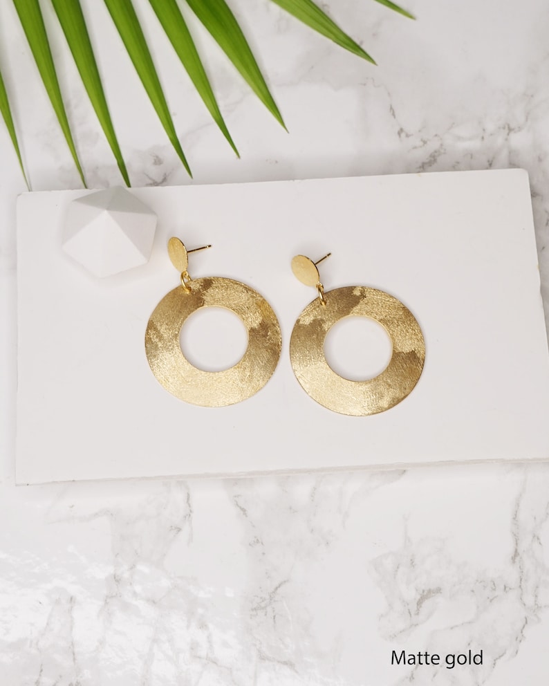 May include: A pair of gold-toned earrings with a circular design. The earrings have a matte finish and are hanging from a small post.