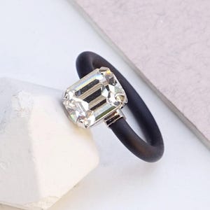 Op de afbeelding: Een ring met een rechthoekige, smaragdgeslepen heldere kristal in een zilverkleurige metalen zetting, bevestigd aan een gladde, matzwarte ringband. De tekst CLEAR CRYSTAL staat in de rechterbenedenhoek van de afbeelding.