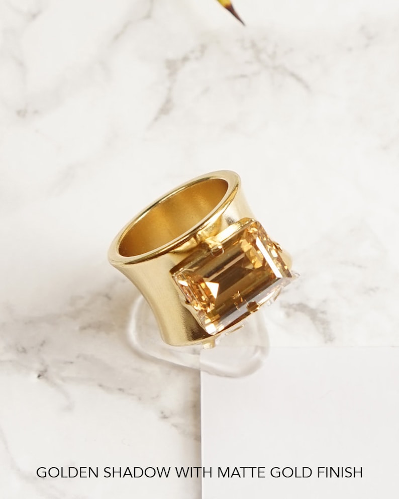 May include: A gold ring with a large, rectangular, light brown gemstone. The ring has a wide band with a matte finish.