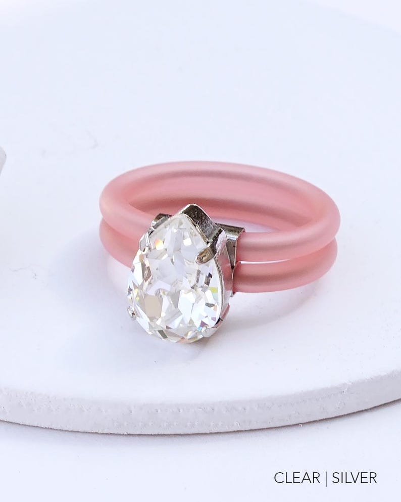 May include: A pink silicone ring with a clear pear-shaped crystal. The ring is double-banded and has a silver setting for the crystal. CLEAR | SILVER