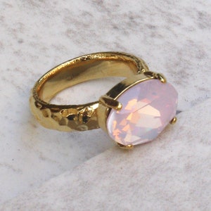 May include: A gold-toned ring featuring a faceted, oval-shaped pink opal stone set in a four-prong setting. The band of the ring has a textured, hammered finish. The text "Pink Opal" is printed in the bottom left corner of the image.